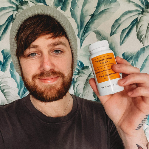 A man with a beanie smiling and holding a Hey Nutrition Turmeric Extract Complex supplement bottle in front of a leafy wallpaper background.