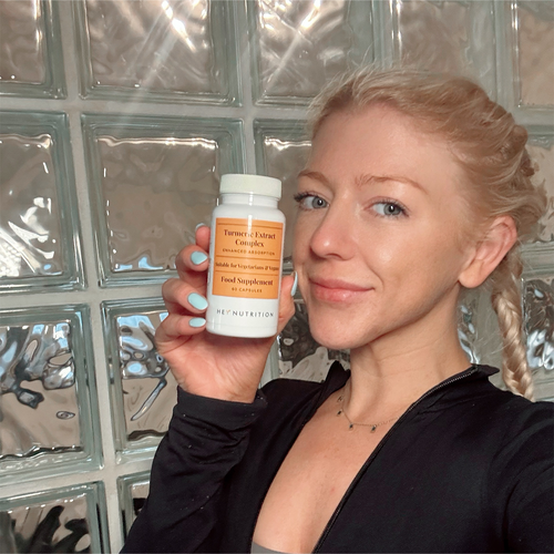 A woman with braided blonde hair smiling and holding a Hey Nutrition Turmeric Extract Complex supplement bottle, standing in front of a glass block wall.