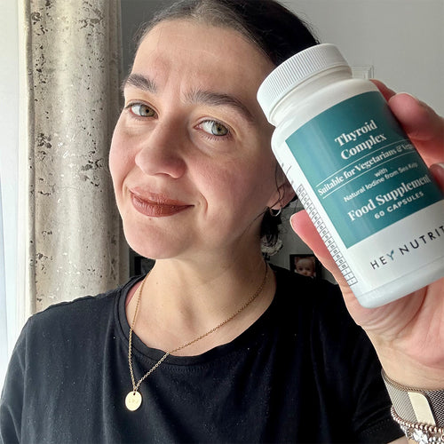 A woman with dark hair in a black top smiling and holding a bottle of Hey Nutrition Thyroid Complex close to her face, highlighting the supplement for thyroid health and balance.