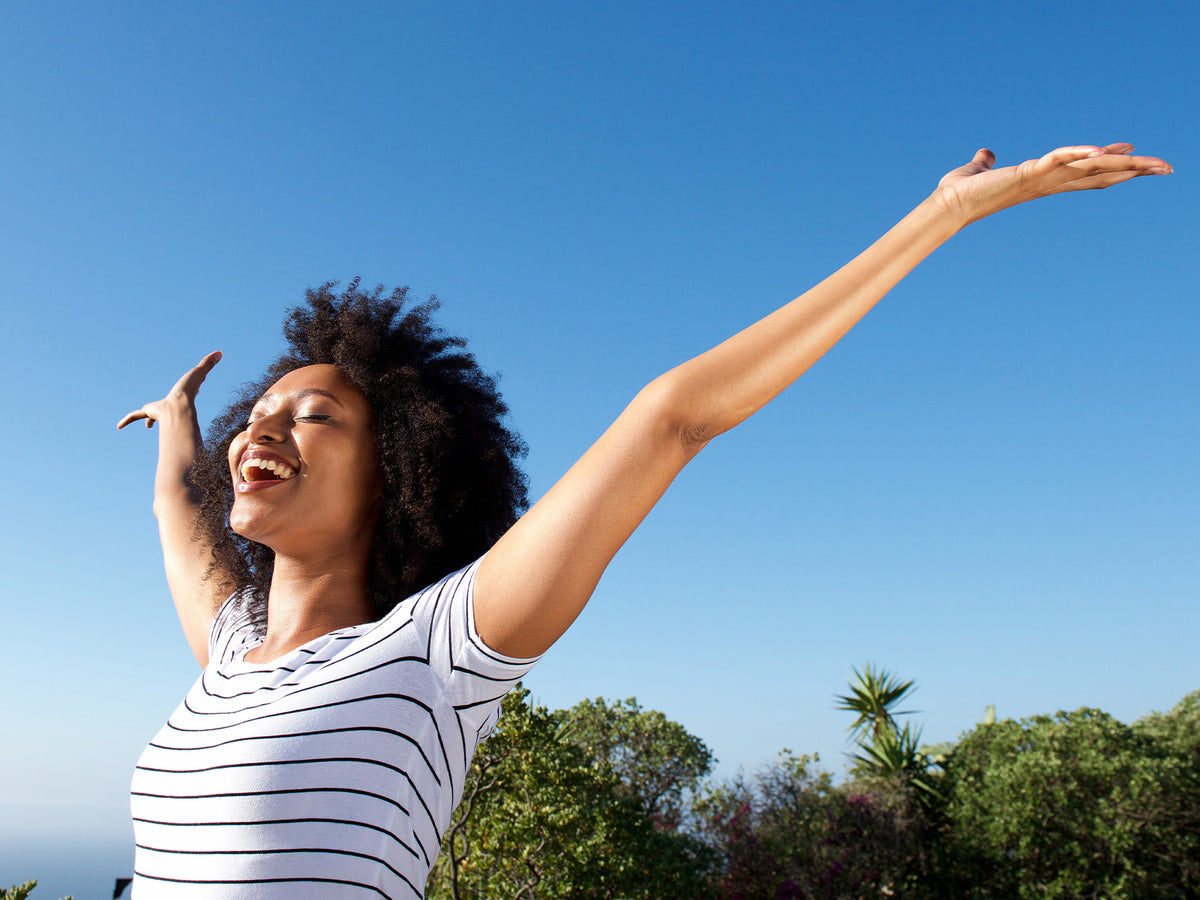 A joyful woman with curly hair wearing a striped t-shirt smiles with her arms stretched wide under a bright blue sky, symbolizing energy and vitality.