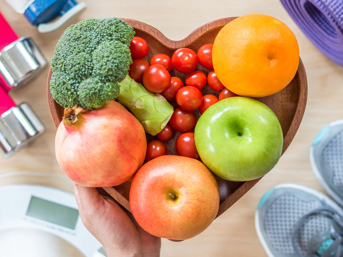 A heart-shaped wooden bowl filled with fresh fruits and vegetables including broccoli, pomegranate, apples, orange, and cherry tomatoes, surrounded by fitness items like dumbbells and a yoga mat.