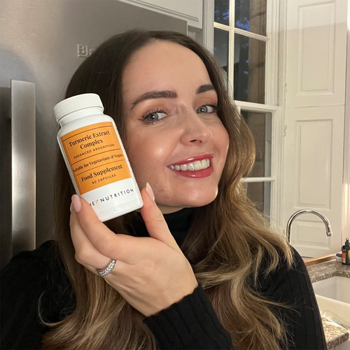 A woman with long brown hair smiling and holding a Hey Nutrition Turmeric Extract Complex supplement bottle close to her face, photographed indoors.