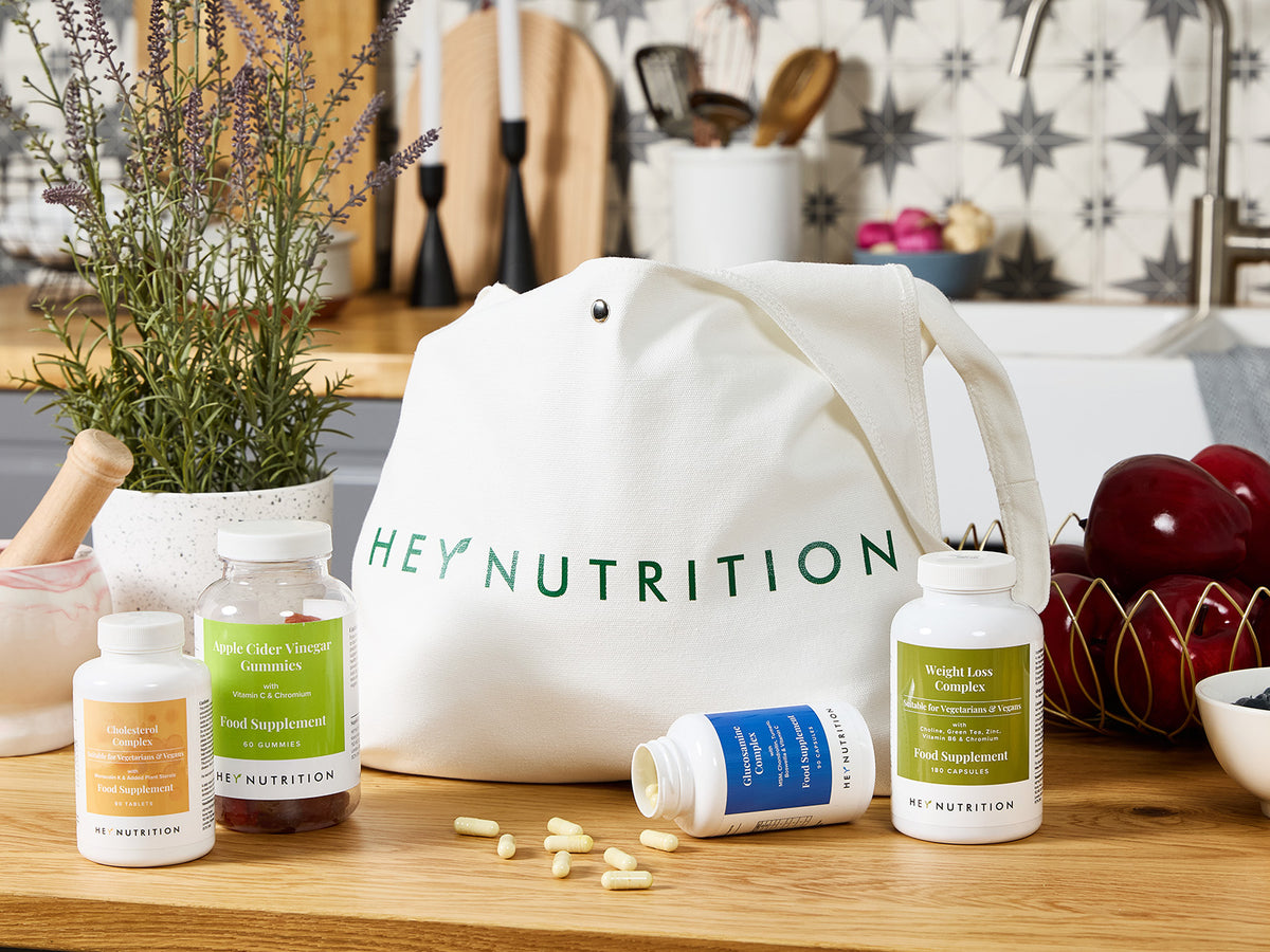 A kitchen counter scene featuring Hey Nutrition supplements arranged around a white tote bag with the brand logo, creating a lifestyle display with natural decor elements.