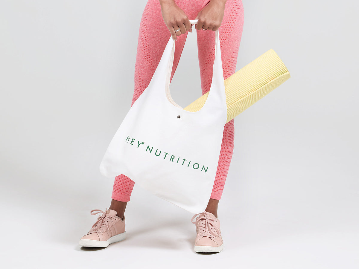 A person in pink activewear holding a Hey Nutrition tote bag with a yellow yoga mat inside, highlighting convenience and on-the-go health.
