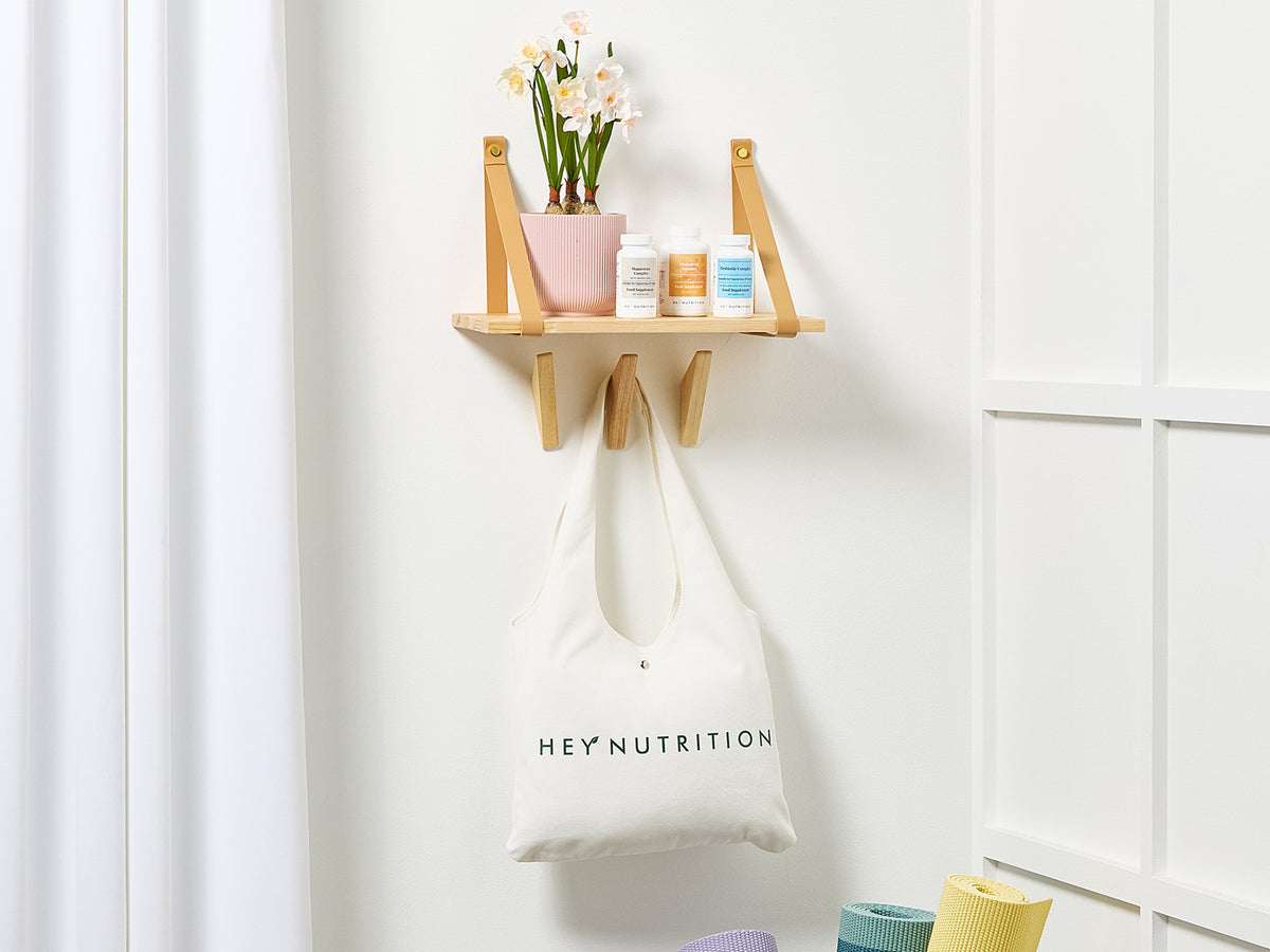 A neatly styled interior with a white Hey Nutrition tote bag hanging under a wooden shelf that displays supplements and a potted plant, suggesting home wellness and balance.