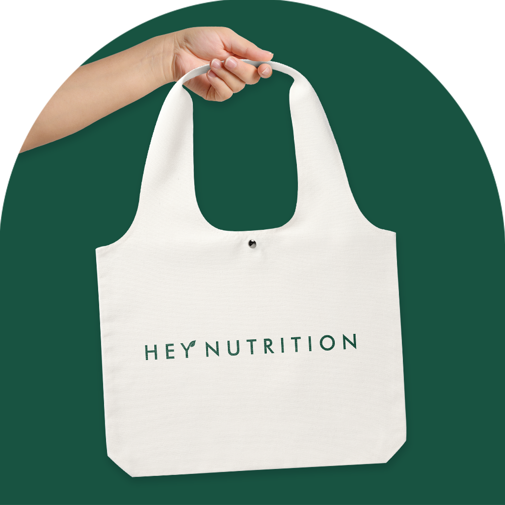 A close-up of a hand holding the minimalist white Hey Nutrition tote bag against a dark green background, emphasizing simplicity and modern branding.