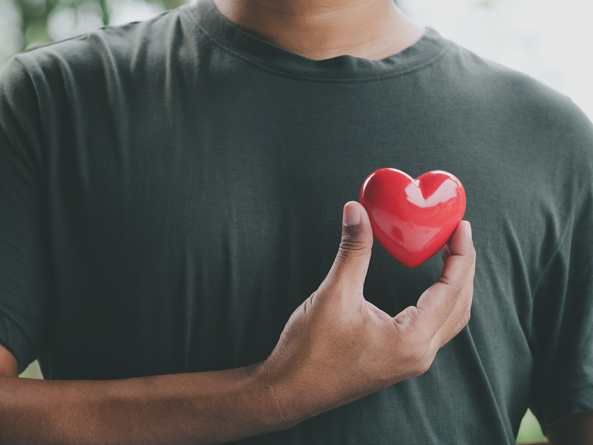 A man in a dark t-shirt holds a bright red heart-shaped object close to his chest, symbolizing cardiovascular health and emotional well-being.