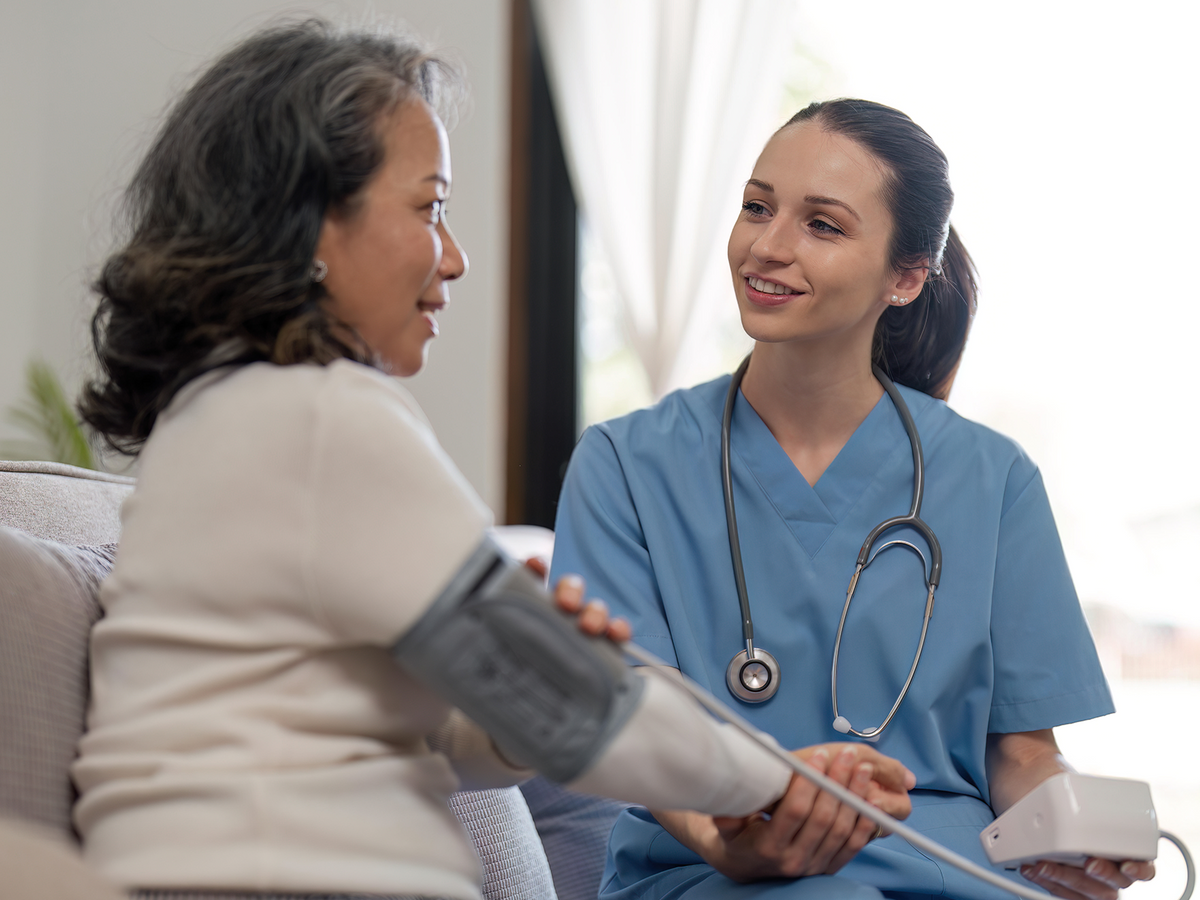 A smiling nurse in blue scrubs measures an older woman’s blood pressure in a home or clinic setting, illustrating professional health monitoring and care.