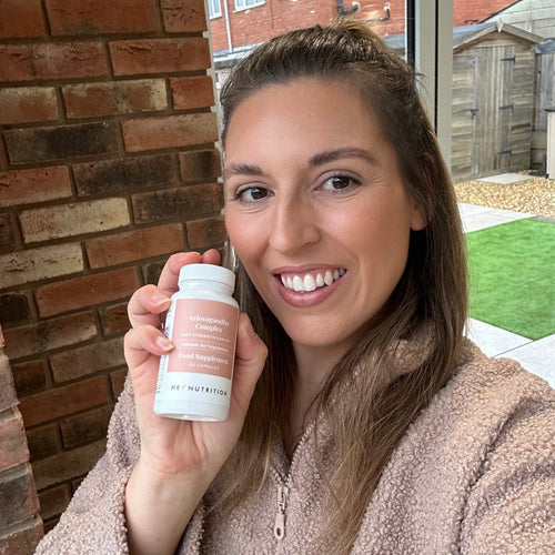 Woman smiling outdoors in front of a brick wall, holding a bottle of Hey Nutrition Ashwagandha Complex.