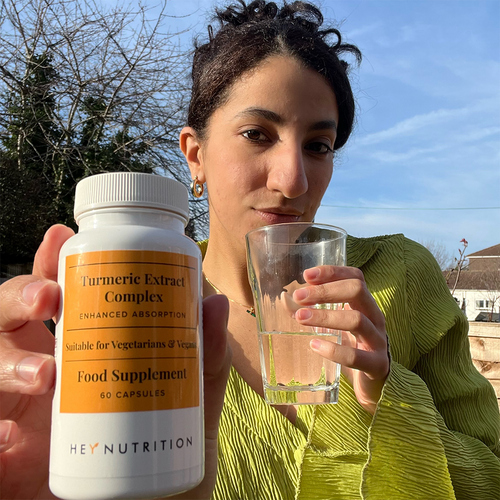 A woman in a green blouse holding a Hey Nutrition Turmeric Extract Complex supplement bottle in one hand and a glass of water in the other, standing outdoors in natural light.