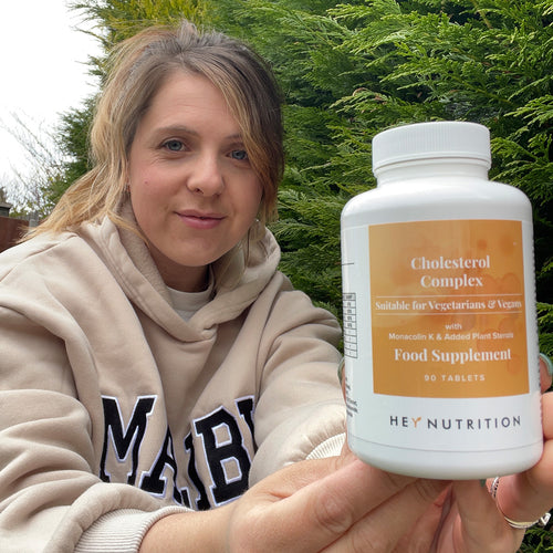 Outdoor shot of a woman in a hoodie seated by evergreens, arm extended to showcase a large Hey Nutrition bottle labeled “Cholesterol Complex — Suitable for Vegetarians & Vegans — with Monacolin K & Added Plant Sterols — Food Supplement — 90 tablets.”